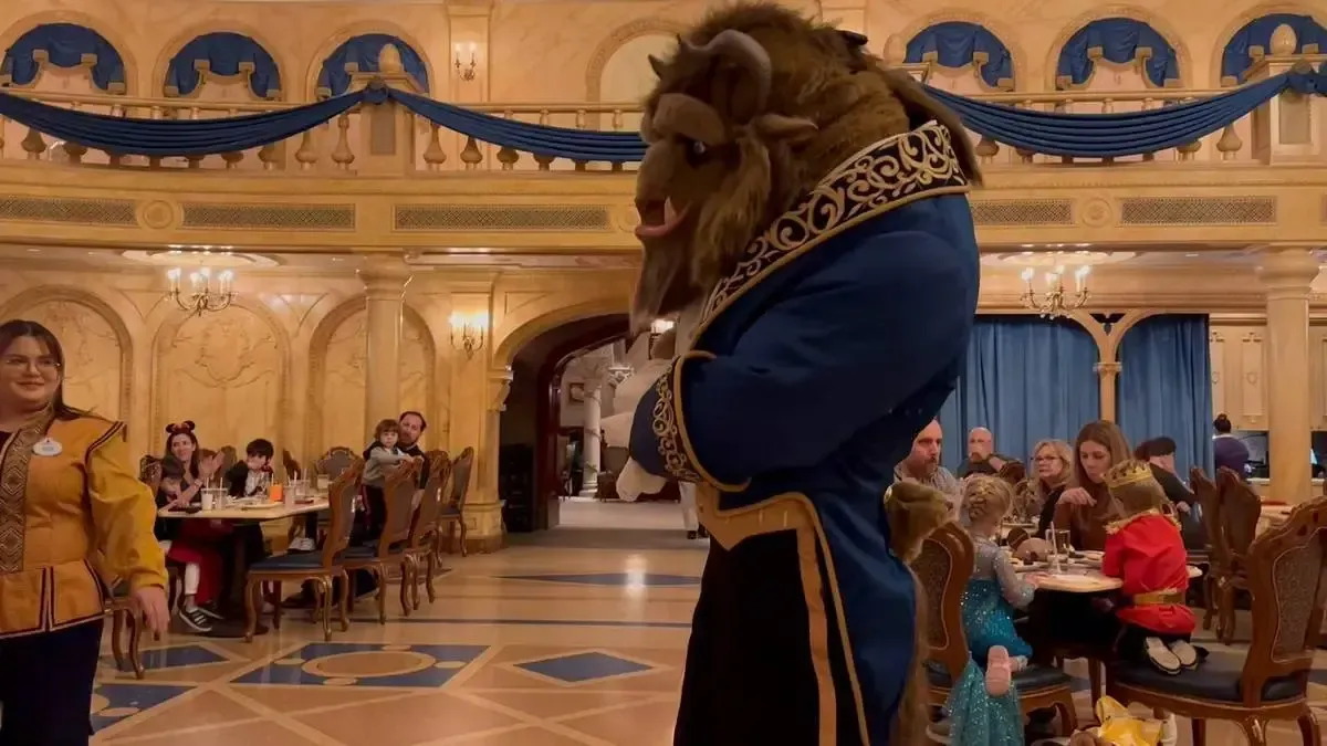 A person in a Beast costume from "Beauty and the Beast" stands in a grand dining room with ornate decor and blue drapes. Diners, including children in costumes, are seated at tables enjoying their meals. A staff member in themed attire walks nearby.