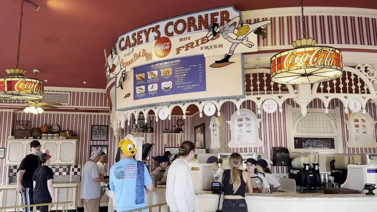 Customers stand at the counter of Casey's Corner, a vintage-style eatery with striped red walls and Coca-Cola themed decor. The menu above features hot dogs and fries, and a playful cartoon character is part of the signage.