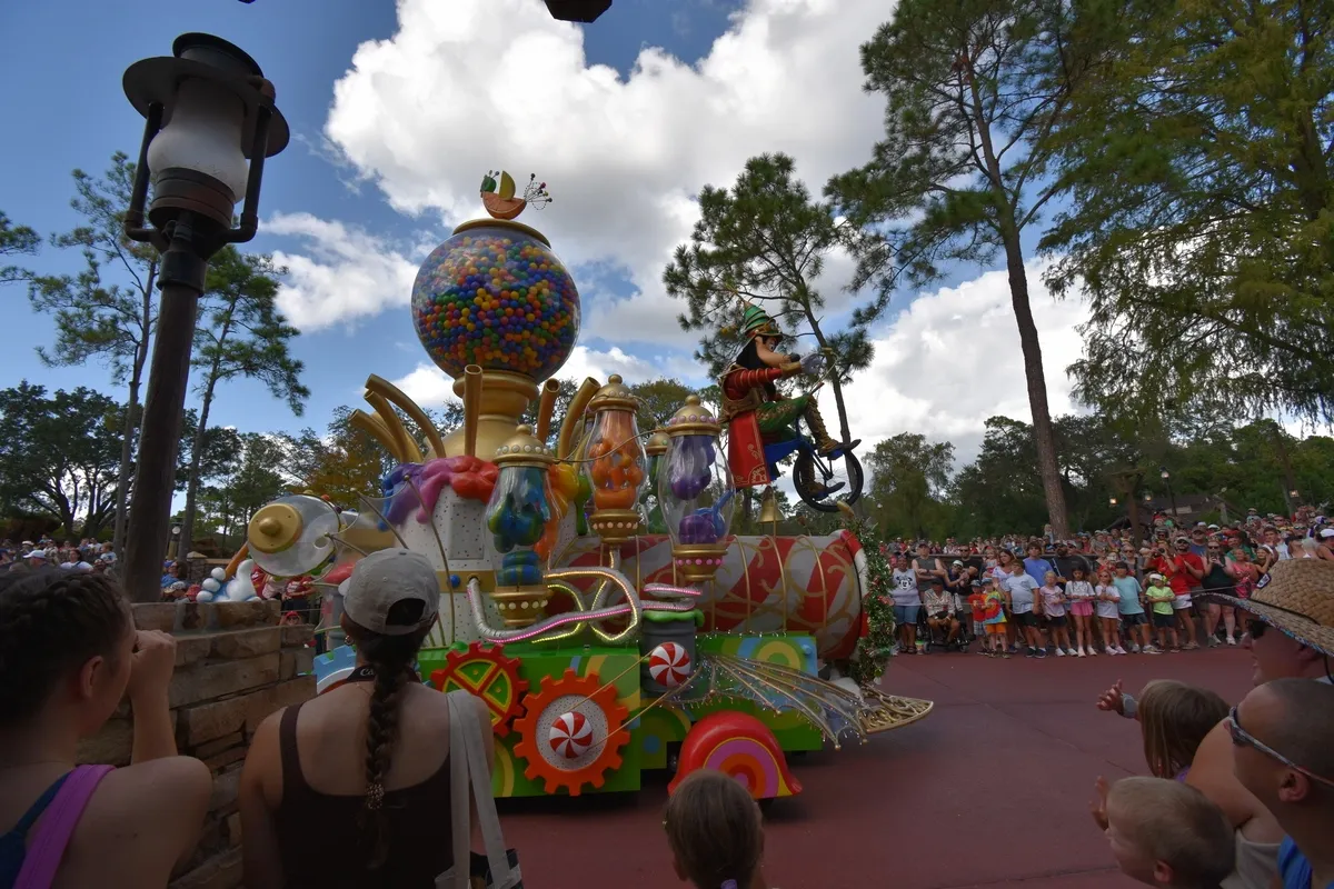 A colorful parade float featuring a whimsical design with a large gumball machine and various candy decorations moves along a crowded street. A character in a green hat rides a unicycle atop the float, entertaining the gathered crowd of onlookers, including children and adults, under a bright, partly cloudy sky.