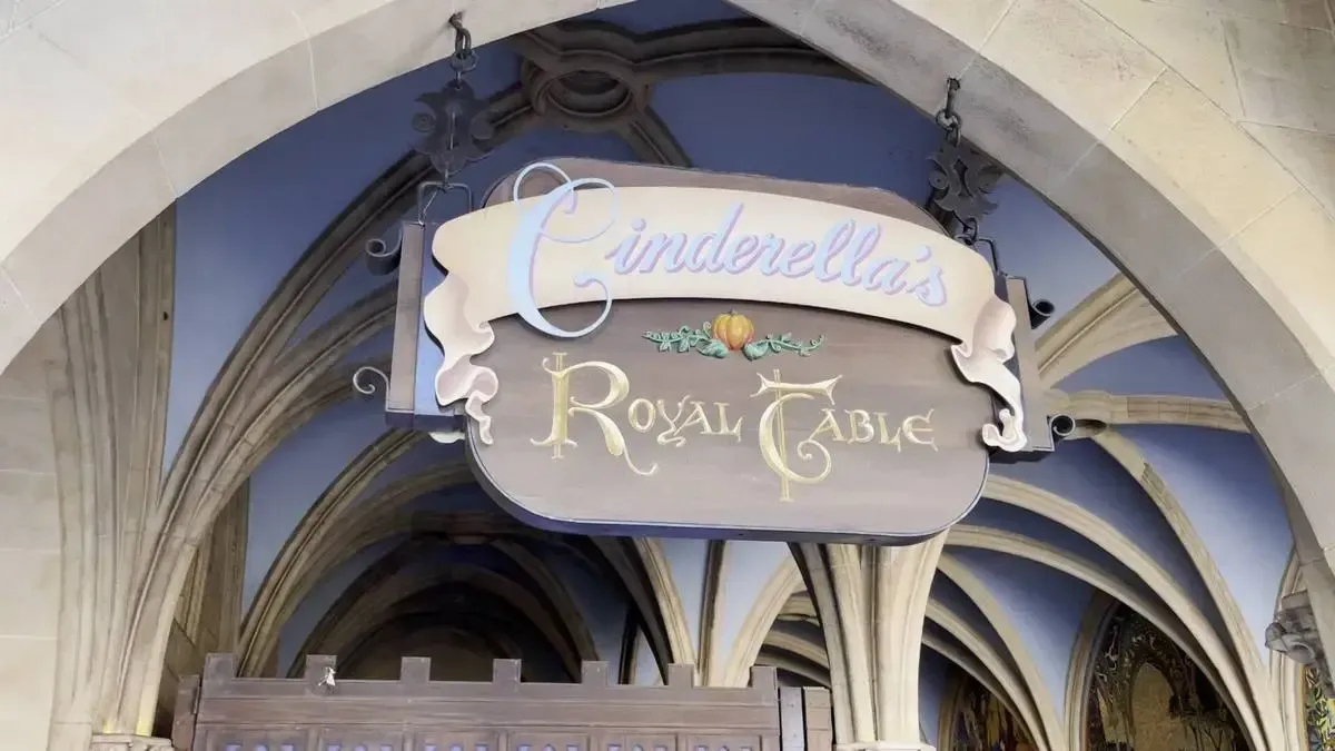 A decorative sign for "Cinderella's Royal Table" hangs under an arched stone entrance, featuring elegant script and a small pumpkin design, set against a background of ornate architectural details.