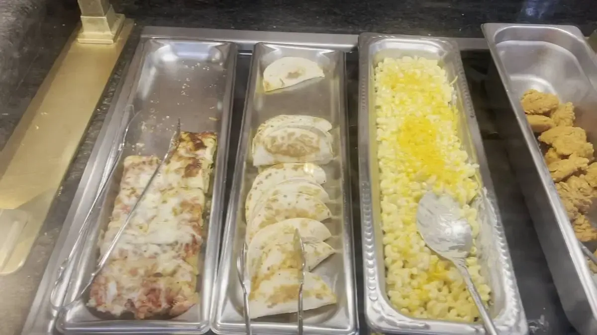 A buffet display features several food trays: one with pizza slices, another with folded quesadillas, a third with macaroni and cheese topped with corn, and a fourth containing pieces of fried chicken.