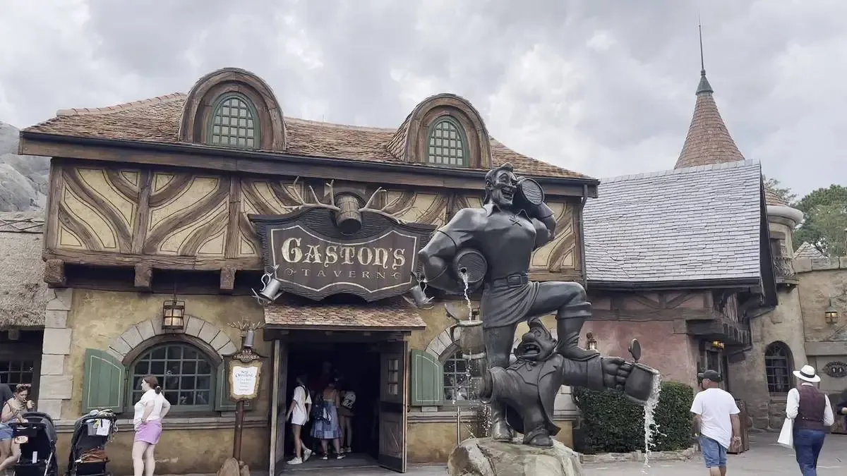 A rustic building with a sign reading "Gaston's Tavern" features a prominent statue of Gaston from Beauty and the Beast holding barrels. The structure has a charming, fairy-tale design with rounded windows and a thatched roof. Visitors are seen entering and walking around the area.