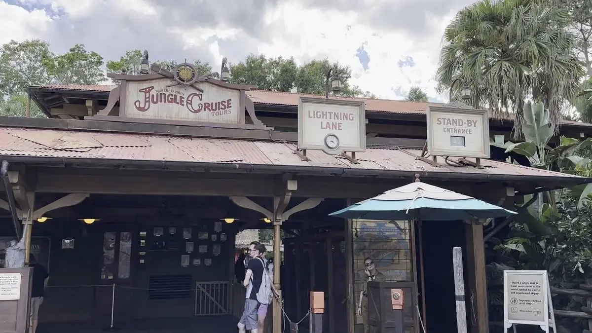 The entrance to the Jungle Cruise attraction features a rustic wooden sign, with separate lanes marked "Lightning Lane" and "Stand-by Entrance." Lush greenery surrounds the building, and a person stands near the entrance under a blue umbrella.