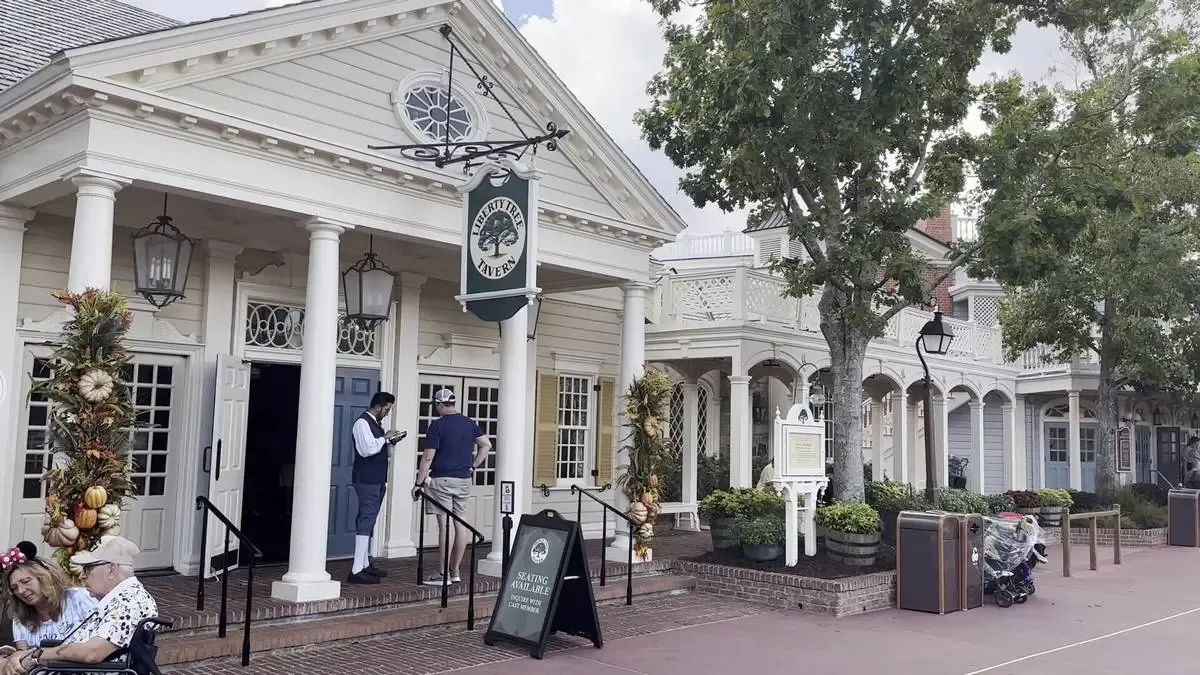 A colonial-style building with white pillars and a sign reading "Liberty Tree Tavern" hangs outside. A person in historical attire stands near the entrance, while a couple sits nearby under autumn-themed decorations. Trees and additional buildings line the brick walkway.
