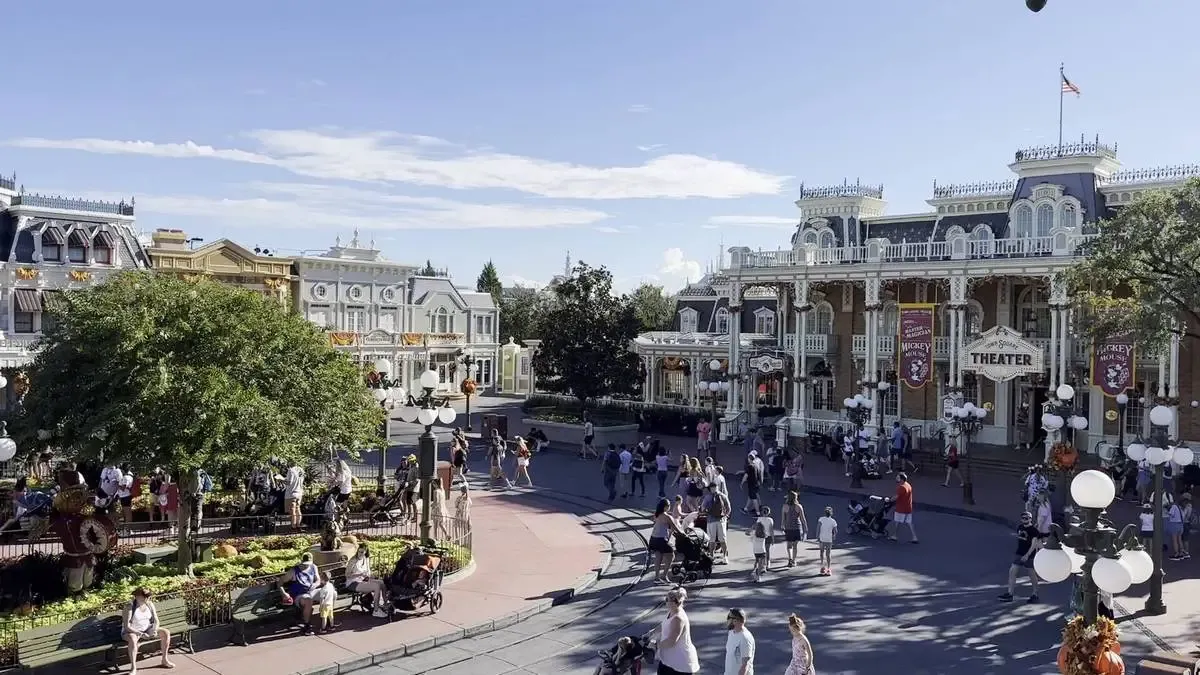 Aerial scene at Magic Kingdom shows a bustling street with people strolling, some with strollers, in front of ornate, Victorian-style buildings adorned with colorful banners. 