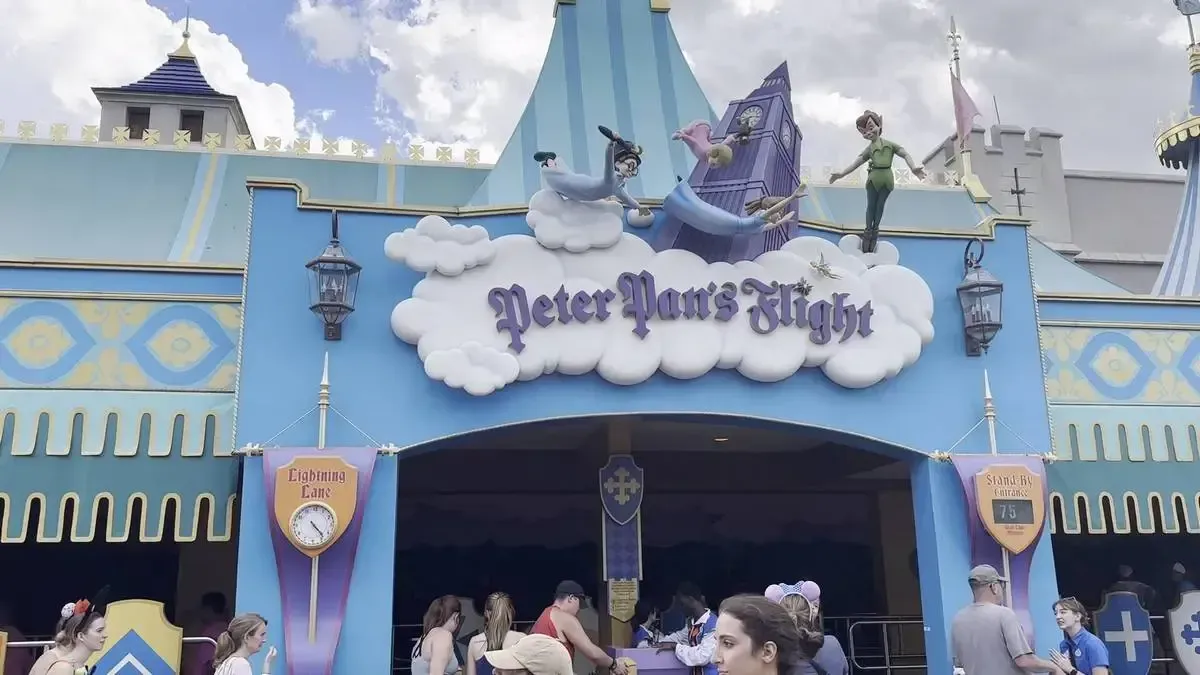 The entrance to "Peter Pan's Flight" features a whimsical design with characters flying over a sign that has the shape of a cloud. The vibrant blue facade is adorned with lanterns, and people are gathered below, waiting in line.