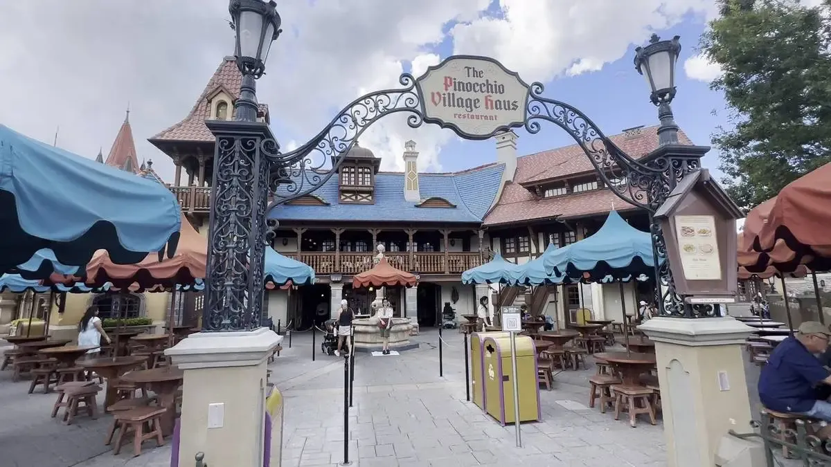 The entrance to The Pinocchio Village Haus restaurant features a decorative wrought iron archway with a sign, surrounded by colorful umbrellas covering outdoor wooden tables, against a backdrop of a charming, European style building with a blue roof.