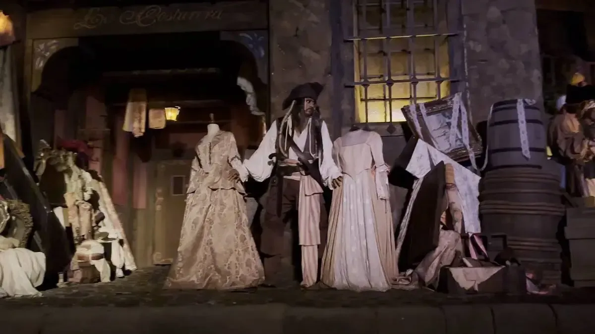 A Jack Sparrow's mannequin stands among various elegant dresses and barrels in a rustic setting, resembling a scene from a themed attraction.