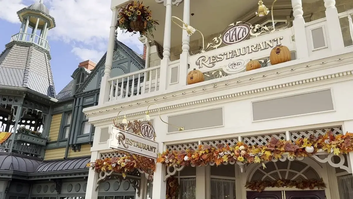 The exterior of the Plaza Restaurant is decorated for fall, featuring pumpkins and garlands of autumn leaves. The building has a charming, vintage architectural style, with ornate details and a sign prominently displaying "Plaza Restaurant." 