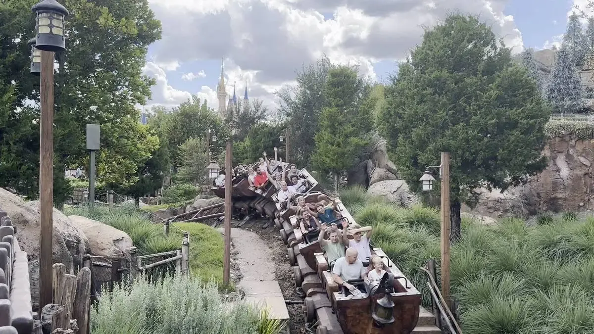 A roller coaster with several passengers moves through a lush, green landscape under a partly cloudy sky, with a castle visible in the distant background.