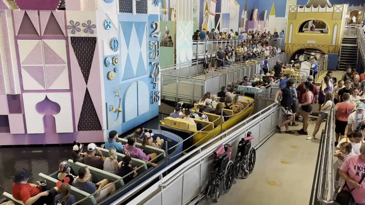 People seated in colorful boats on a water ride inside a theme park, with vibrant geometric decorations on the walls. A crowd waits in line beside the ride, and several wheelchairs are parked nearby.