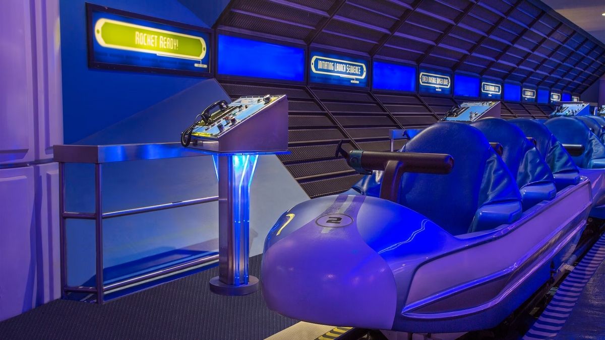 A futuristic ride vehicle with blue seats is positioned in an area with space decoration. Illuminated signs on the wall display instructions, while a control panel with buttons stands nearby, enhancing the high-tech atmosphere.