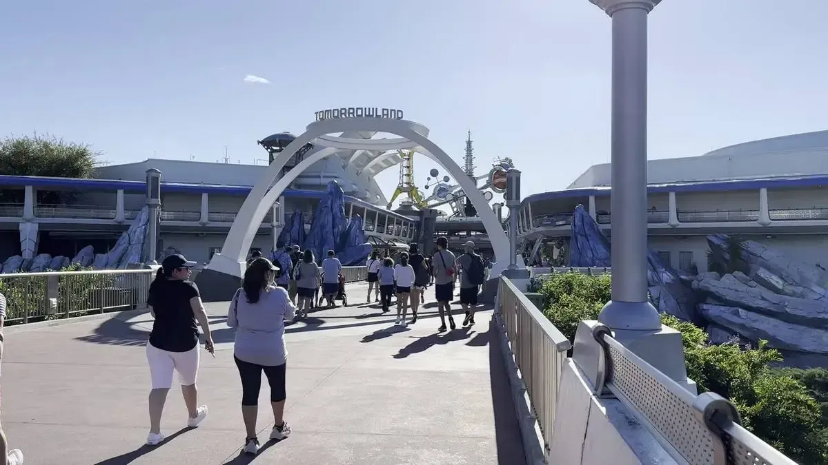 A group of people walk toward the futuristic entrance of Tomorrowland, featuring a large arch and sleek architecture. Blue and white structures and a clear sky create a vibrant atmosphere, while greenery lines the pathway.