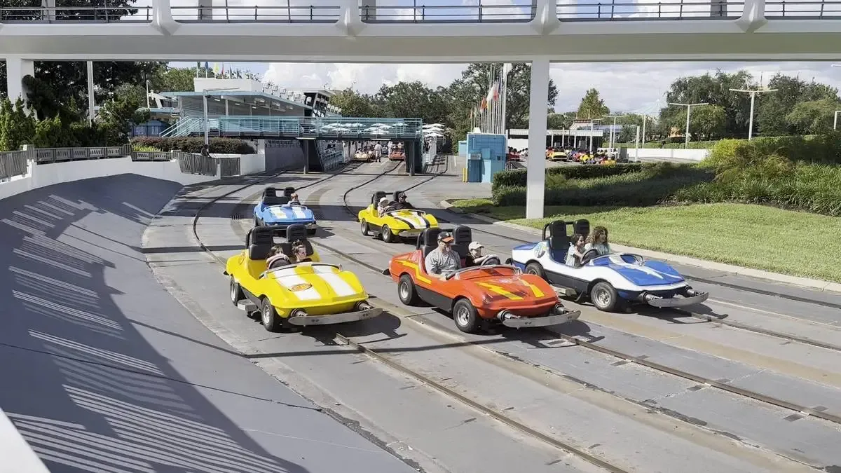 Colorful go-karts with drivers navigate a winding track under a pedestrian bridge, surrounded by greenery and a clear sky.