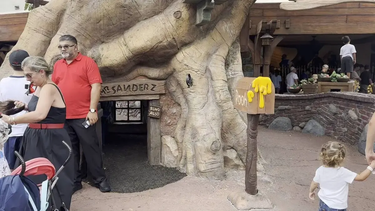 A whimsical tree structure resembling a large trunk with a small door labeled "Mr. Sanders" is surrounded by people at an amusement park. A mailbox labeled "Pooh" with yellow paint dripping from it stands nearby.
