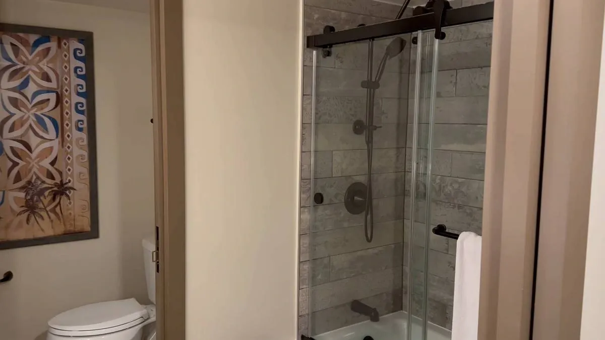 A modern bathroom features a glass-enclosed shower with gray tile walls. To the left, a toilet is partially visible, accompanied by a decorative artwork with a tropical design.