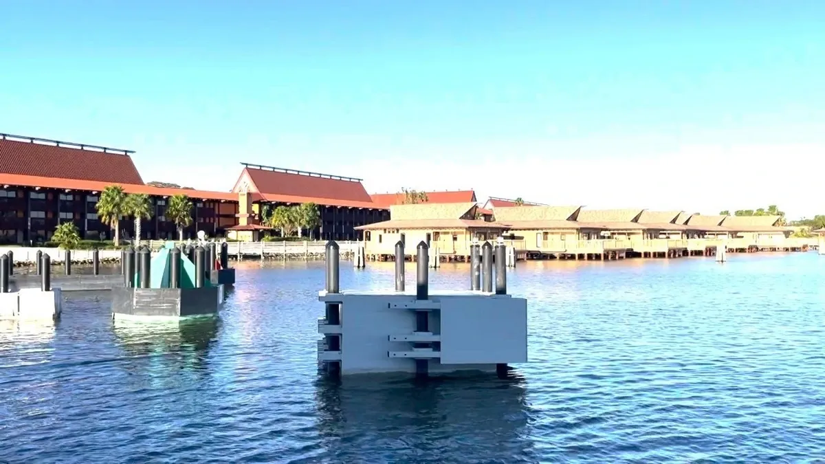 A calm waterfront scene features construction structures in the foreground, partially submerged in the water. In the background, a resort with red-roofed buildings and palm trees lines the shore under a clear blue sky.