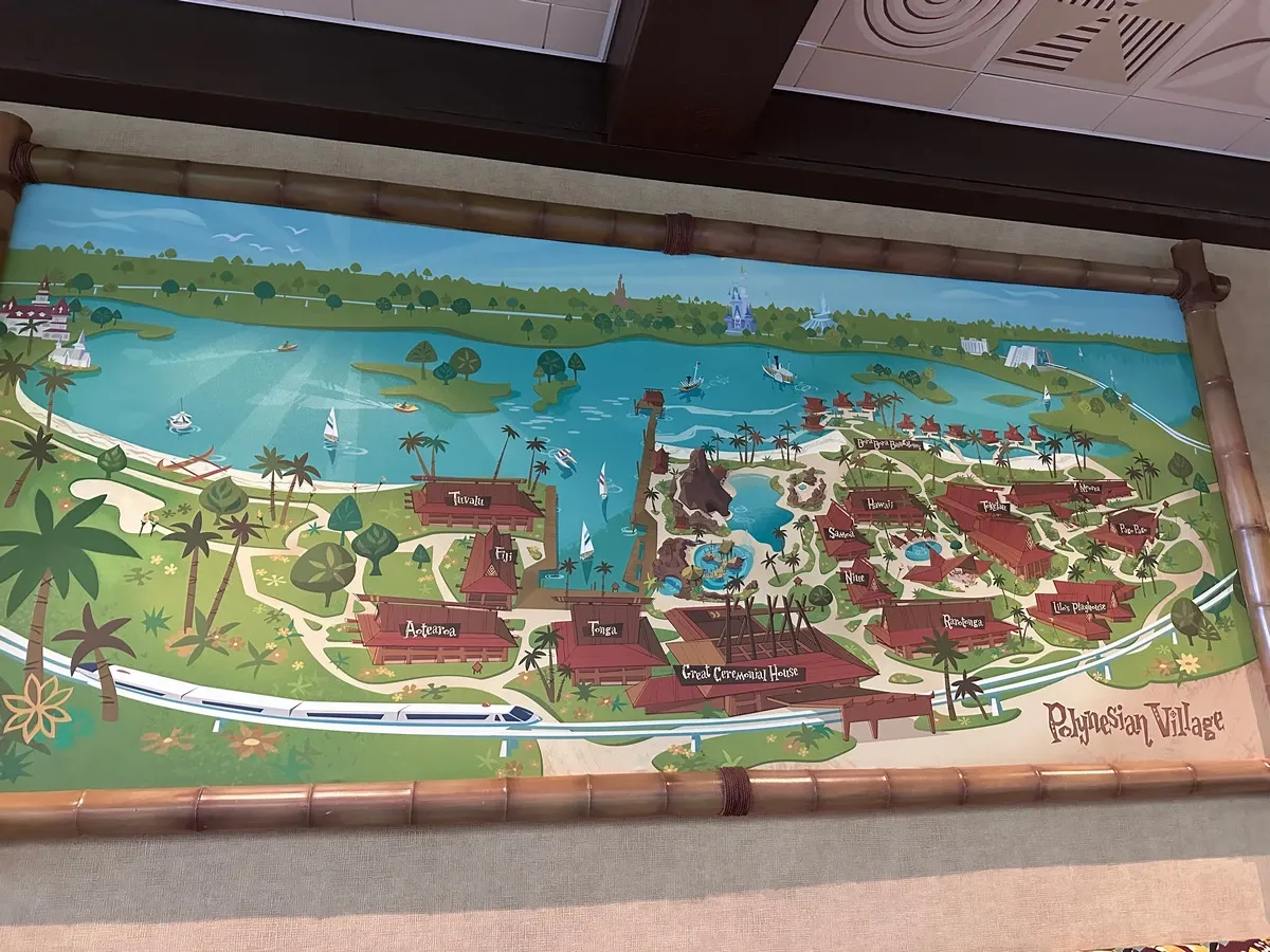 A colorful illustrated map of the Polynesian Village Resort, featuring labeled areas like Tuvalu, Fiji, and Hawaii. The map shows a lagoon with sailboats, tropical landscaping, and a monorail track. A bamboo frame surrounds the map, adding to the tropical theme.