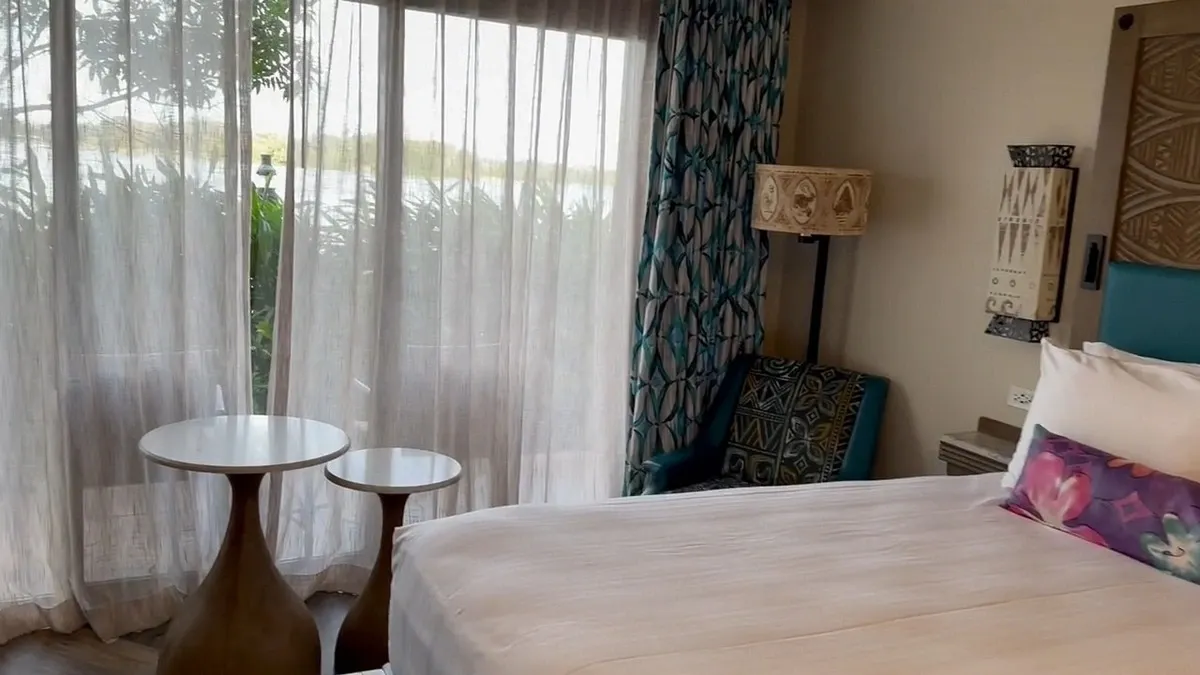 A hotel room featuring a bed next to sheer curtains allowing natural light to filter in. Two small round tables stand by the window, and a patterned armchair is positioned beside a decorative lamp. Lush greenery is visible outside.