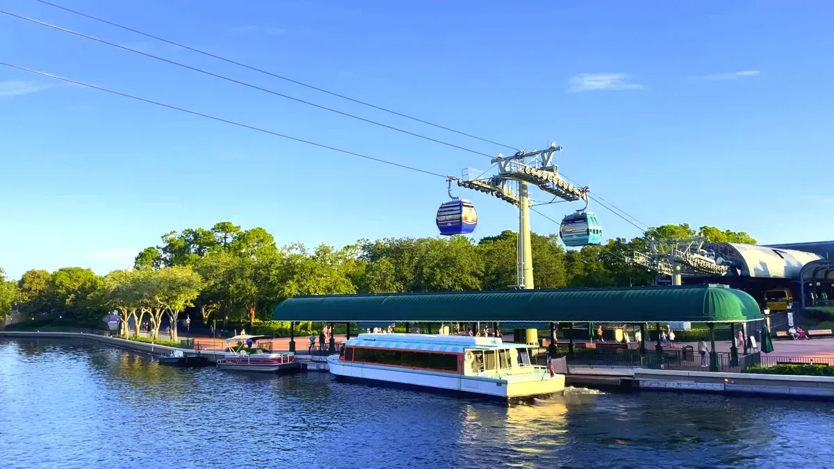 A scenic view of a waterfront with a green-roofed dock and a boat moored alongside, set against a backdrop of lush trees. Above, colorful gondolas glide along cables, part of a cable car system. The sky is clear and blue, enhancing the tranquil atmosphere.