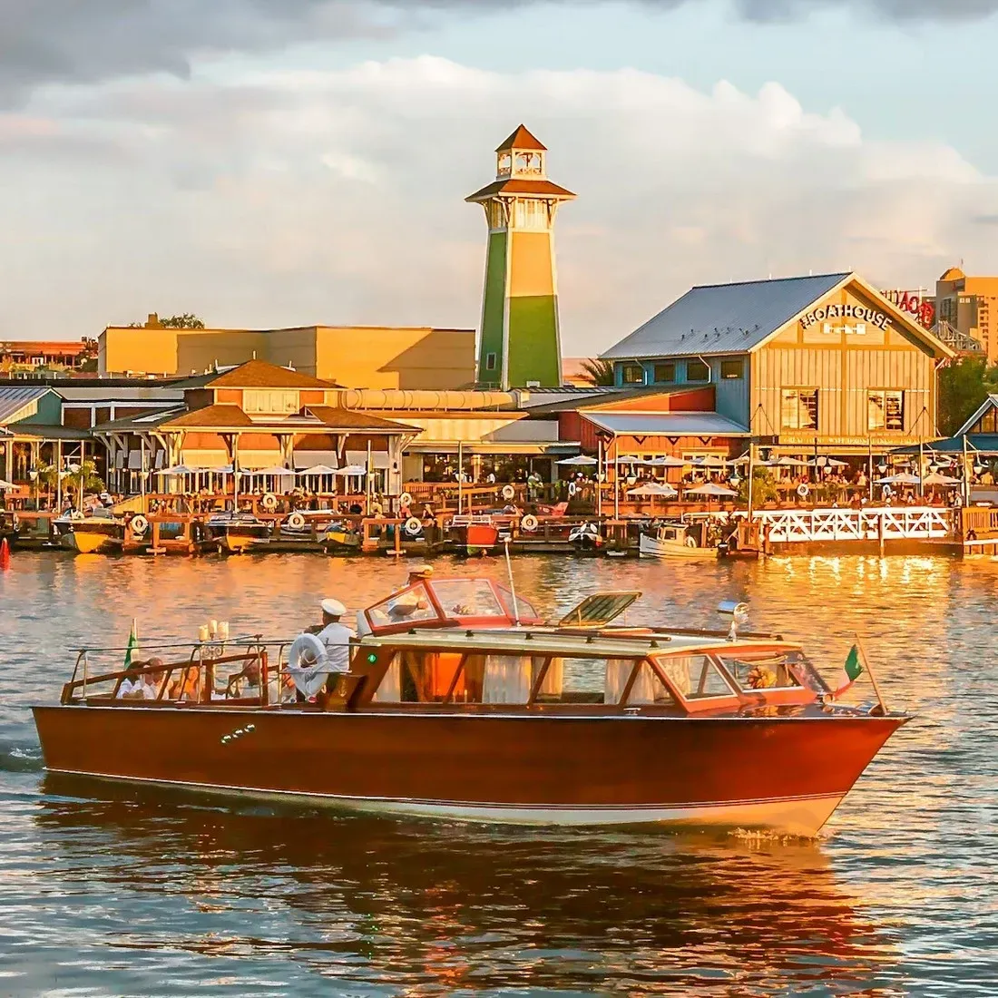 A classic wooden boat cruises on a sunlit waterway, with a lively waterfront scene in the background featuring a lighthouse, bustling restaurants, and colorful buildings.