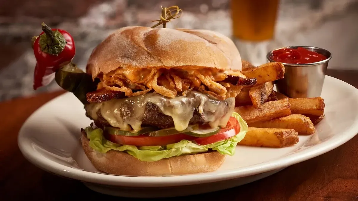 A juicy burger loaded with melted cheese, crispy fried onions, lettuce, and tomato is served on a fresh bun, accompanied by thick-cut fries and a small cup of ketchup on a white plate.