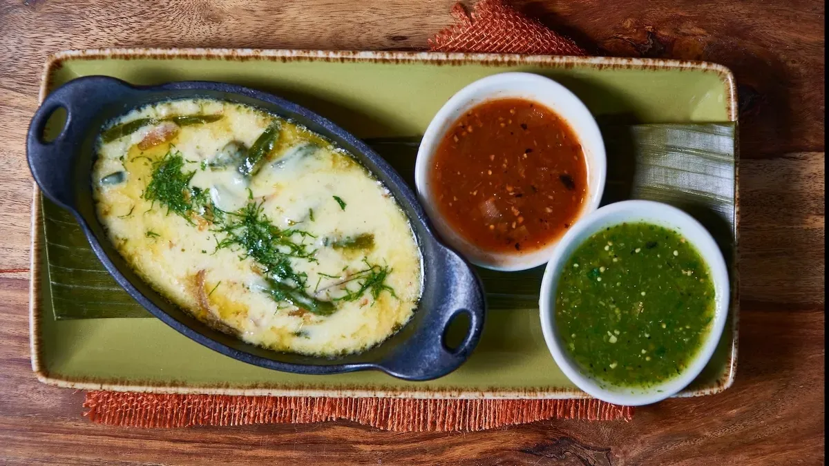 A sizzling dish of melted cheese with herbs and green peppers is served in a black oval skillet, accompanied by two small bowls of red and green salsa on a rectangular green plate.
