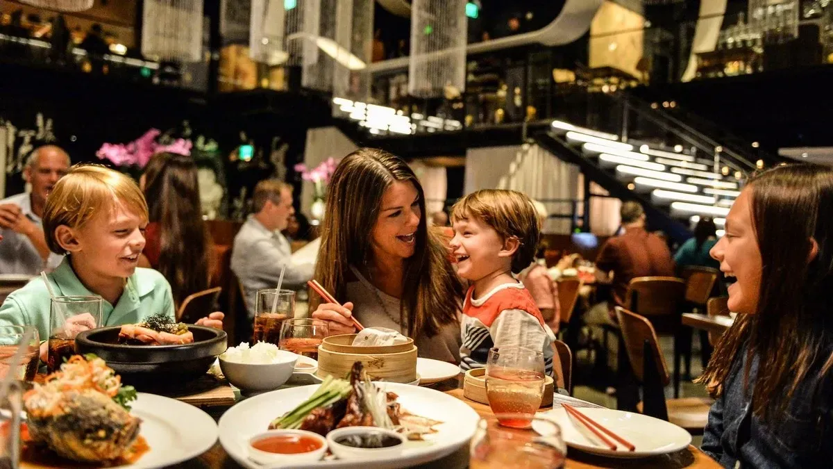 A family is enjoying a meal at a restaurant, with a mother and three children smiling and laughing around a table filled with various dishes. The setting is warmly lit, with a modern interior featuring a staircase and other diners in the background.