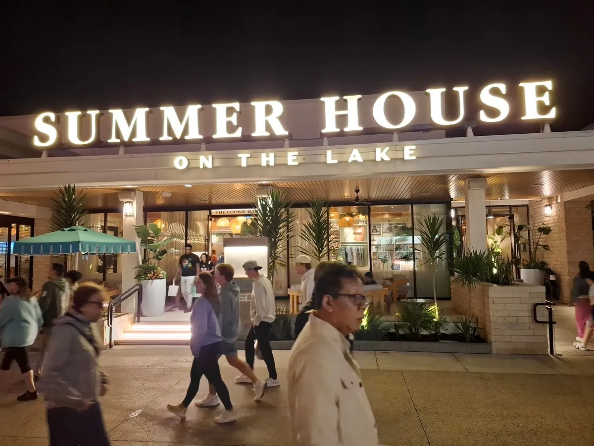People walk past the brightly lit entrance of "Summer House on the Lake" at night, with large potted plants and a teal umbrella adding to the inviting atmosphere.