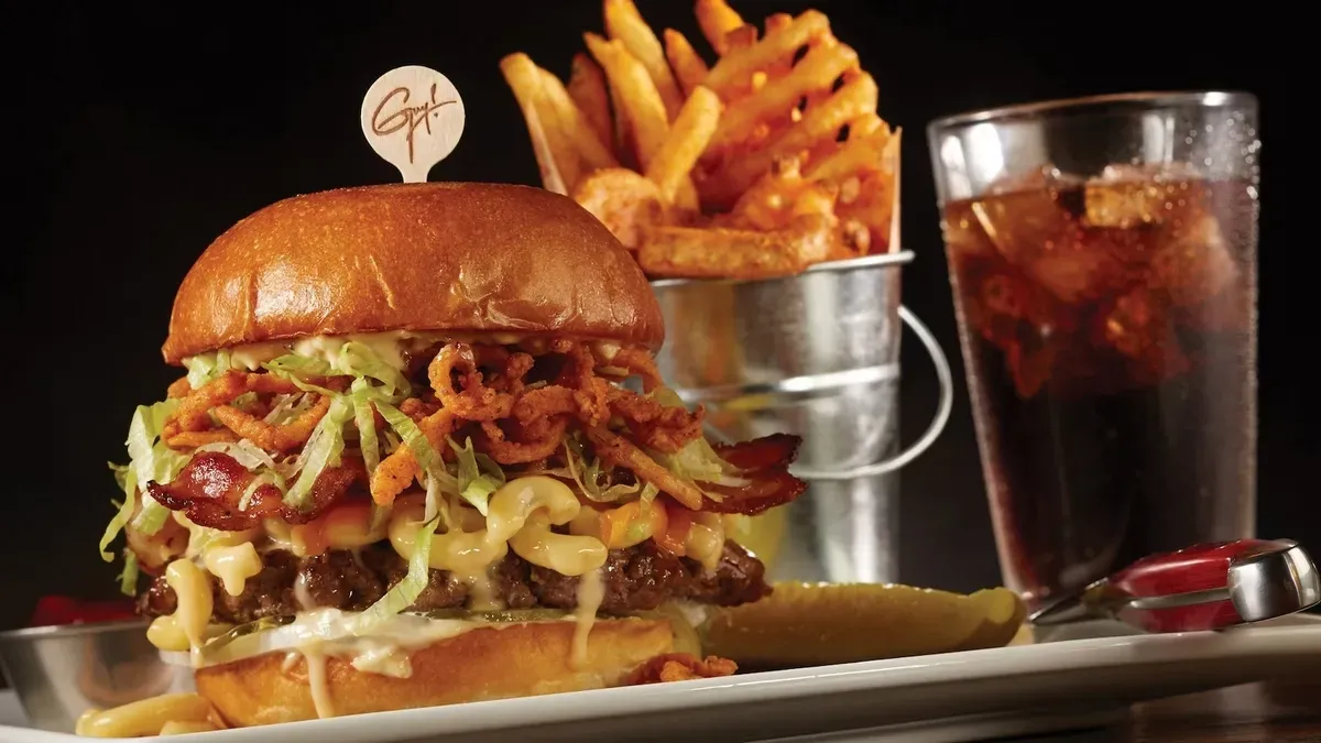 A big burger loaded with bacon, lettuce, crispy onions, and melted cheese sits on a plate with a pickle slice. Next to it, a metal bucket holds golden fries, and a glass of cola with ice completes the meal.