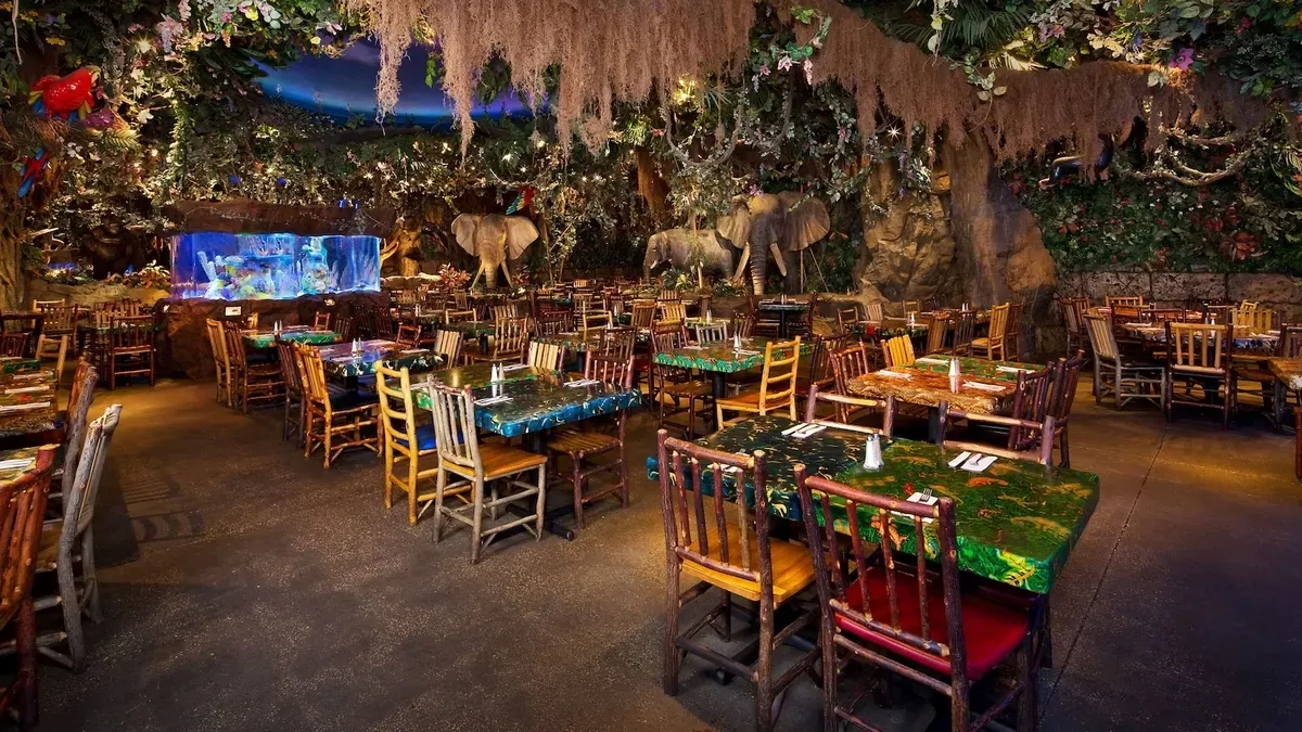 A vibrant rainforest-themed restaurant features colorful tables and wooden chairs, surrounded by lush greenery and hanging vines. An aquarium with bright fish and elephant statues add to the immersive jungle atmosphere.