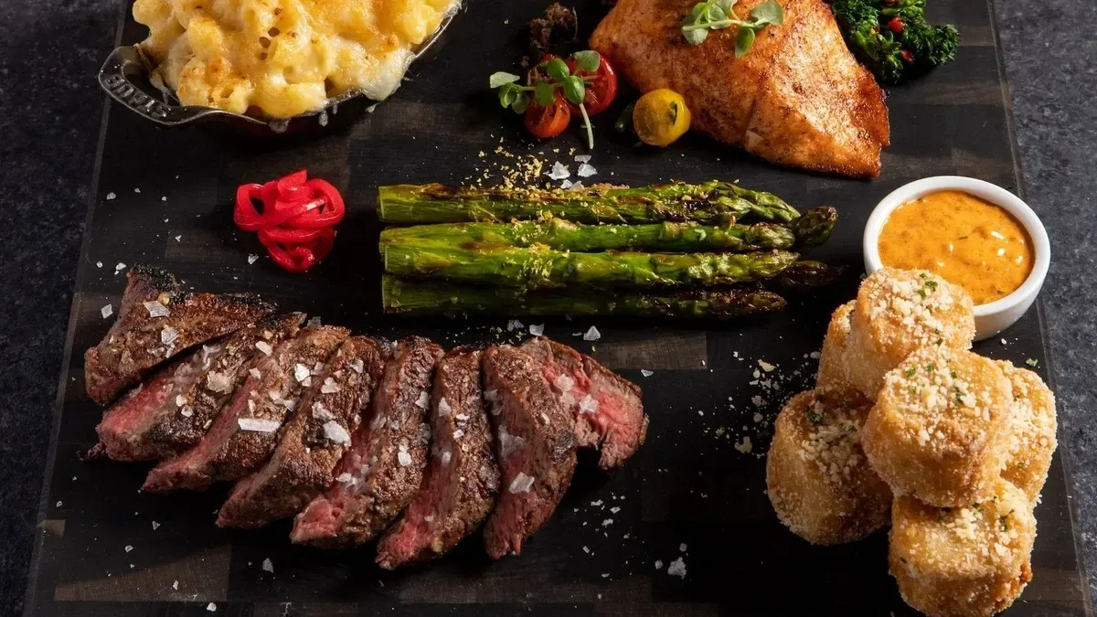 A platter featuring sliced steak topped with coarse salt, grilled asparagus, and a piece of seasoned fish. Accompanying the main dishes are a serving of creamy macaroni and cheese, crispy potato rounds sprinkled with cheese, and a small bowl of orange sauce.