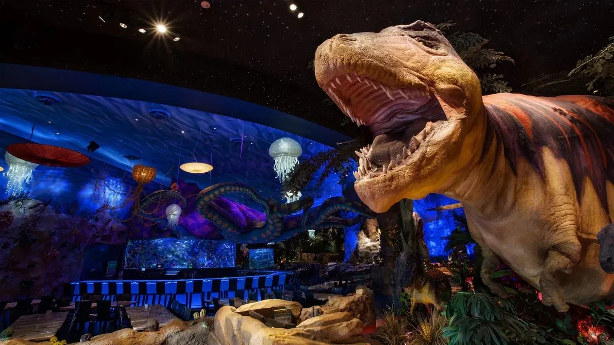 A vibrant, themed restaurant interior featuring a large, lifelike dinosaur model with its mouth open, surrounded by lush greenery. The ceiling is illuminated with blue lighting and decorated with hanging jellyfish and an octopus, creating an underwater ambiance. Tables and chairs are arranged in the foreground, adding to the immersive atmosphere.
