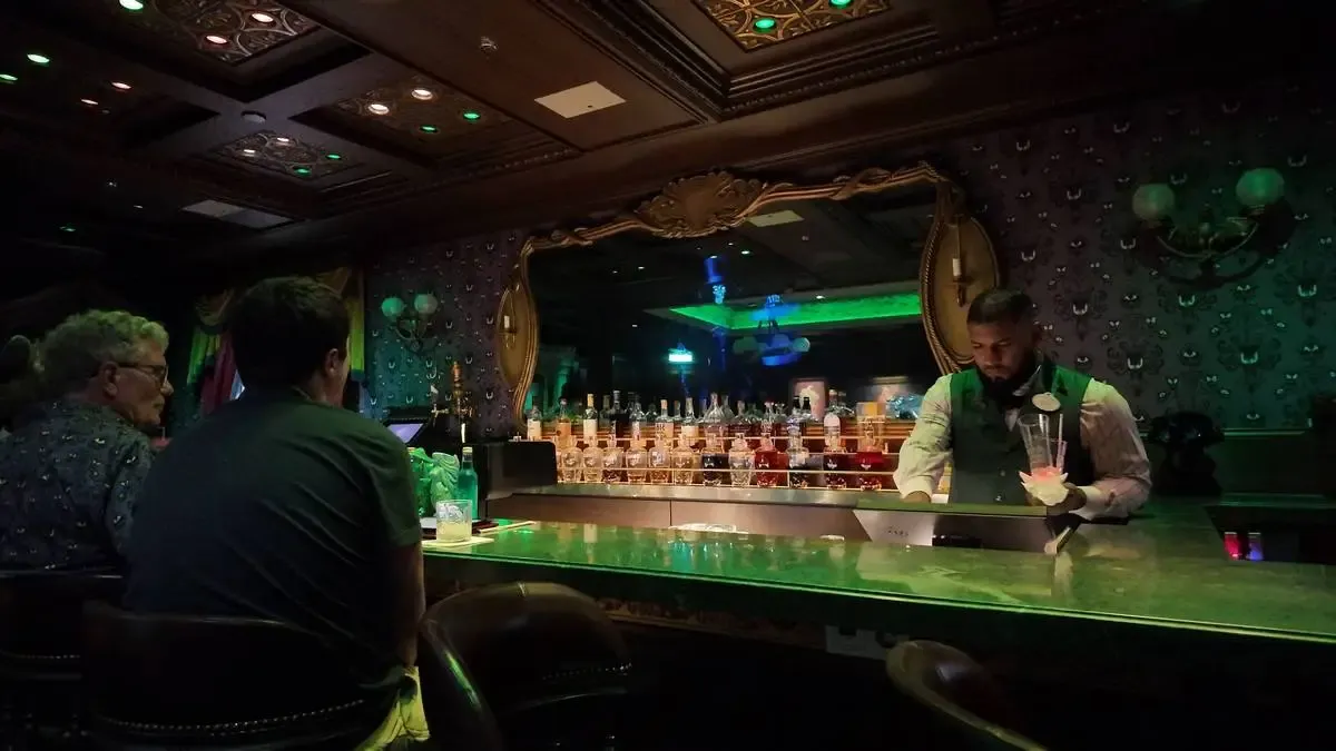 A bartender in a vest prepares a drink at a dimly lit bar, with bottles neatly arranged on the counter. Two patrons sit on bar stools, engaged in conversation, under an ornately decorated ceiling and a large mirror reflecting the ambient glow.