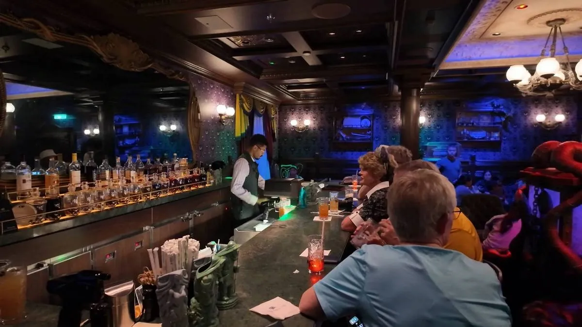 A bartender prepares drinks behind a dimly lit bar, with shelves of various liquor bottles in the background. Several patrons sit at the bar, engaged in conversation, under ornate chandeliers and surrounded by dark, decorative walls.