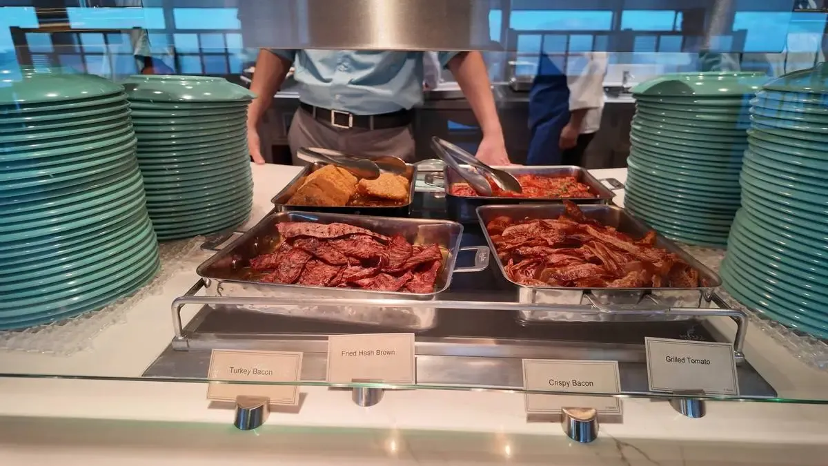 A buffet setup features trays of turkey bacon, fried hash browns, crispy bacon, and grilled tomatoes, with tongs for serving. Stacks of green plates are arranged on either side, and a person in a blue shirt is visible in the background.