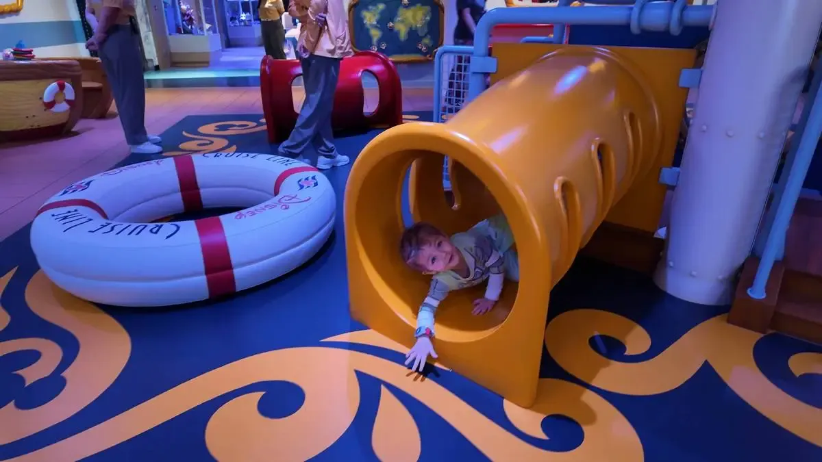 A child is playfully crawling through a bright yellow tunnel in an indoor play area, reaching out with one hand. Nearby, a large inflatable life ring with "Cruise Line" written on it rests on the colorful floor, which features swirling patterns.