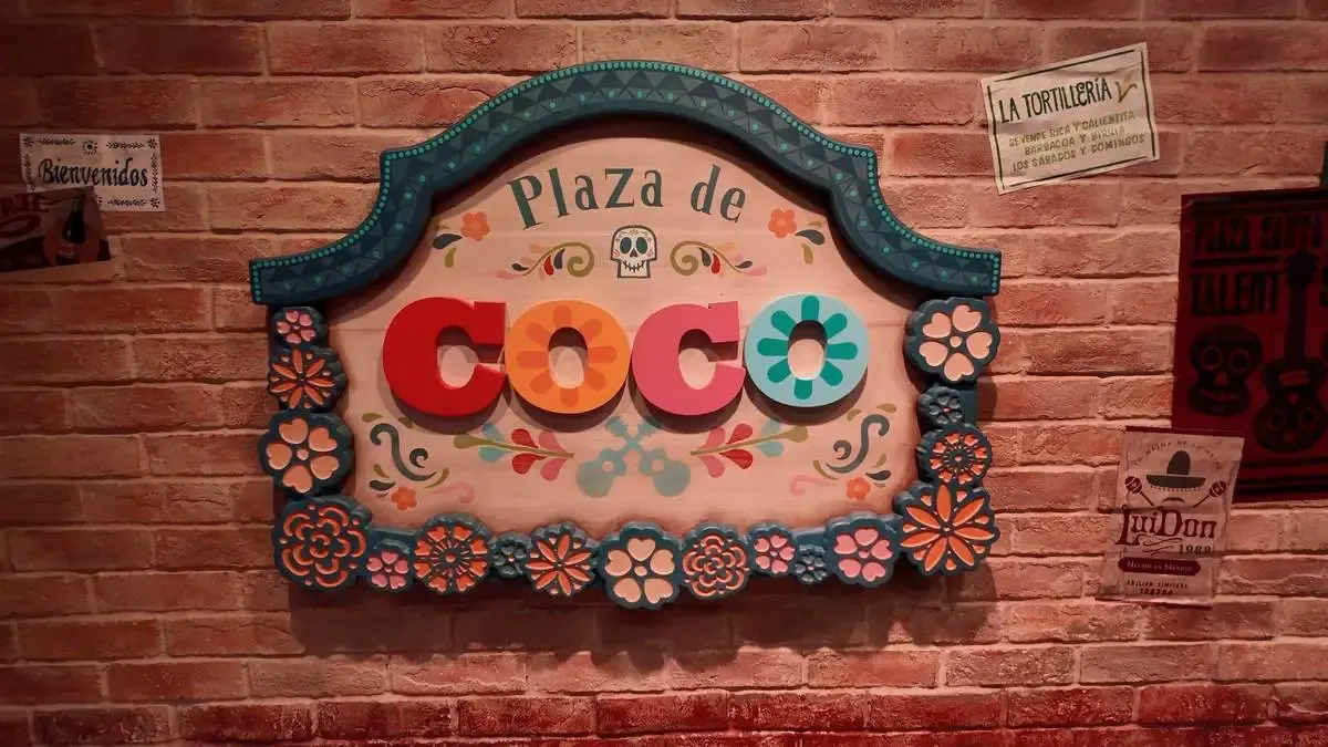 A colorful sign reading "Plaza de Coco" is adorned with vibrant floral decorations and a small skull, set against a brick wall with various posters and signs, creating a festive atmosphere.