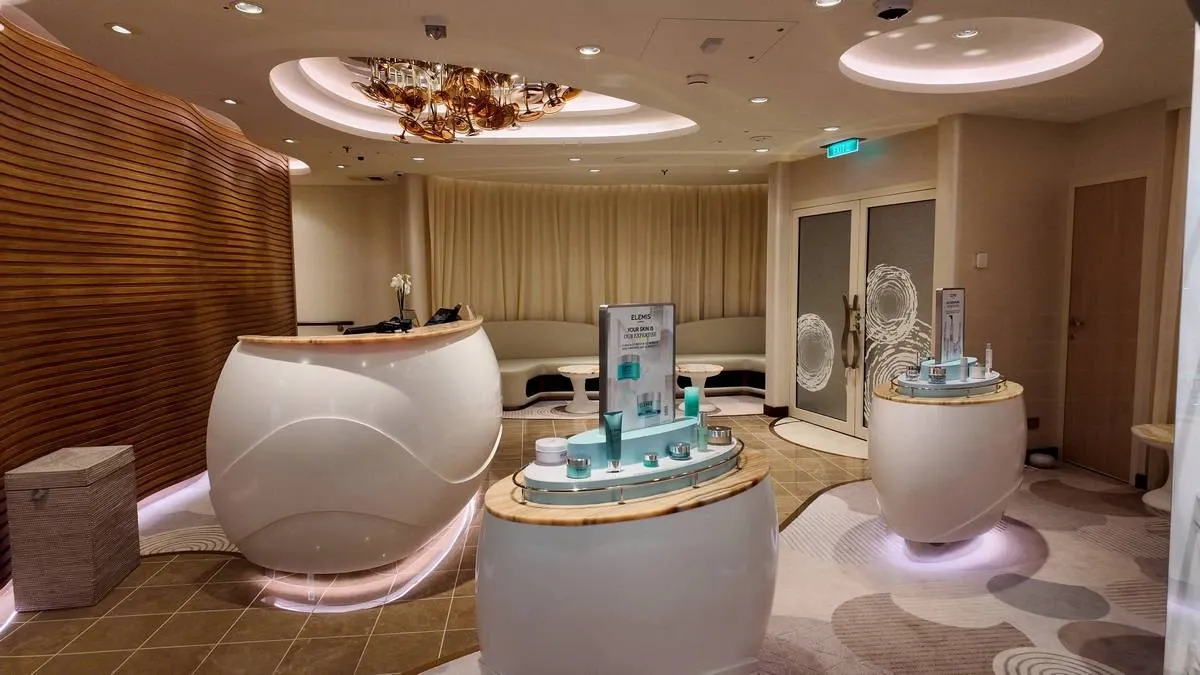 A modern spa reception area features sleek, curved white counters with skincare products displayed. The room has warm wooden accents, soft lighting, and a stylish ceiling fixture, creating a relaxing and luxurious atmosphere.