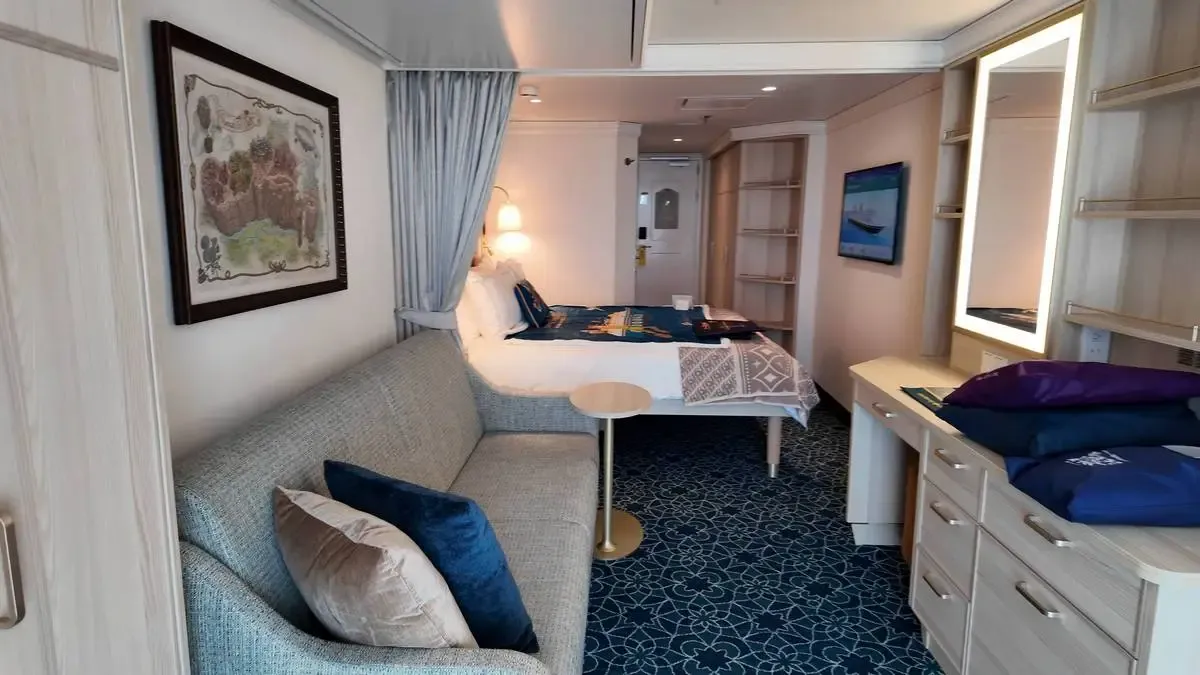 A cozy hotel room features a light gray sofa with blue and beige pillows, a bed with a patterned throw, and a small round table. The room has a decorative wall map, a lit vanity mirror, and a blue patterned carpet, creating a comfortable and inviting atmosphere.