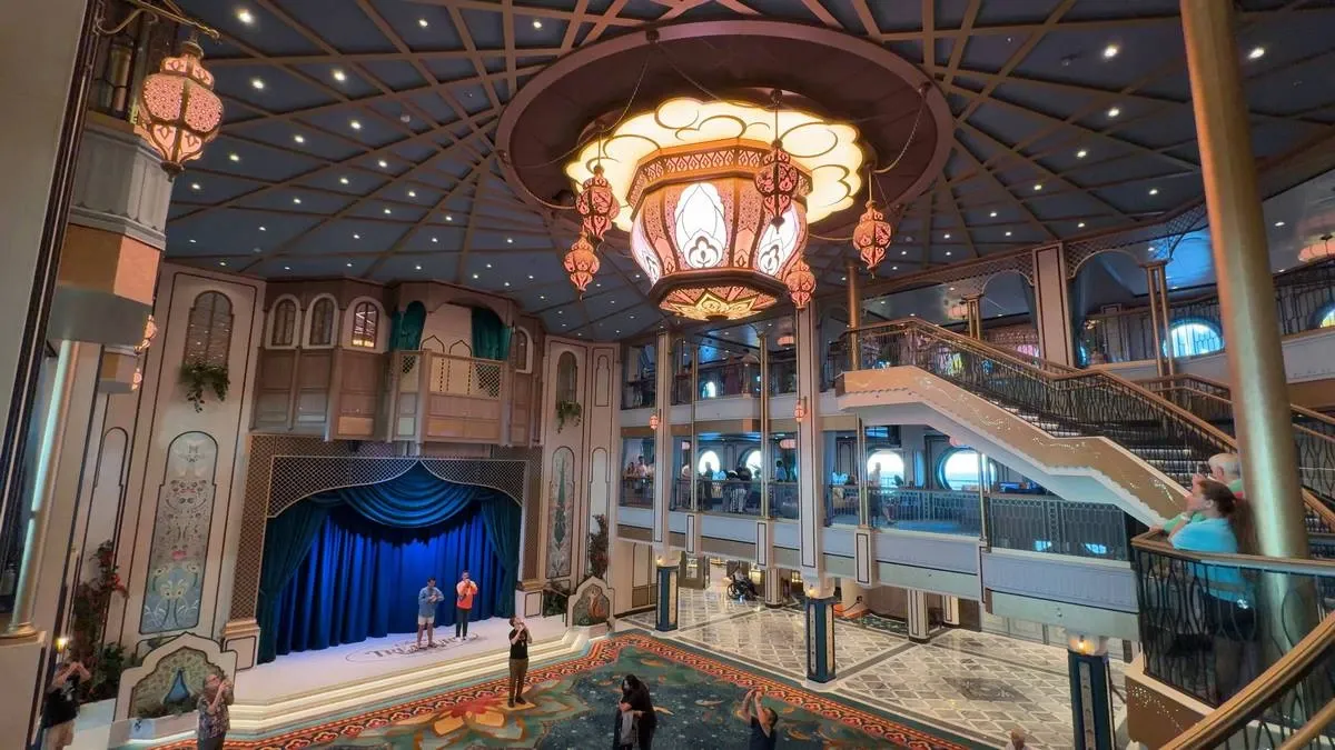 A grand, ornate lobby features a large, decorative chandelier hanging from the ceiling, with intricate patterns and warm lighting. Below, people stand on a patterned floor, some near a small stage with a blue curtain. Elegant staircases with gold accents lead to an upper level, enhancing the opulent atmosphere.