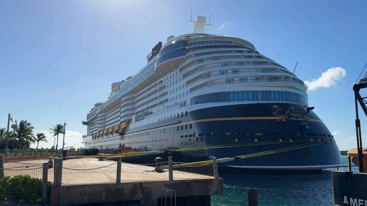 A large cruise ship docked at a sunny port, with its sleek white and blue exterior gleaming under the clear sky. Palm trees and a few flags are visible in the background, adding to the tropical atmosphere.