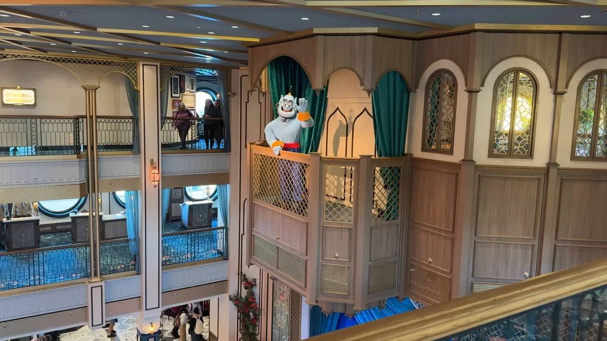 A whimsical character, dressed in a white outfit with red accents, waves from a wooden balcony adorned with green curtains in a multi-level, ornate interior. The space features intricate railings and large round windows, while people stroll on the upper level.