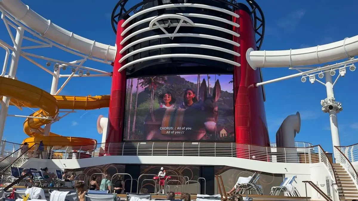 A large outdoor screen on a cruise ship displays a scene from an animated movie. The setting features a bright yellow water slide, lounge chairs, and a clear blue sky, creating a lively atmosphere.