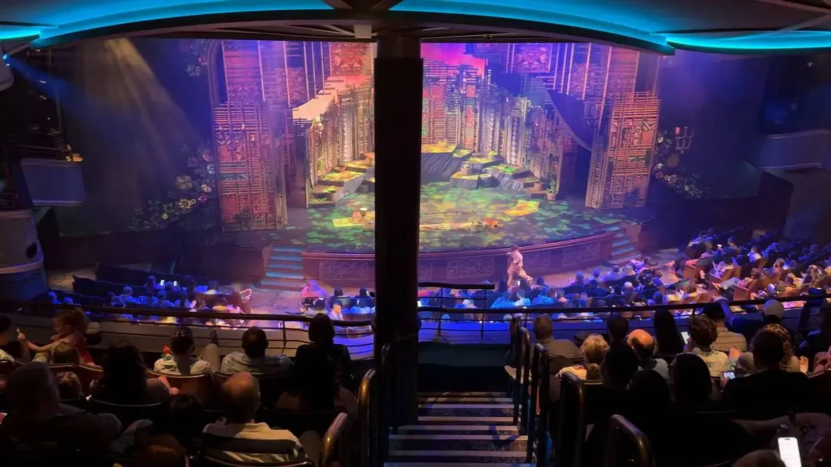 A vibrant theater scene with a packed audience watching a colorful stage set featuring intricate designs and bright lighting. The setting includes a backdrop of vertical structures, possibly representing a cityscape, with an actor visible in front. The atmosphere is lively and engaging, with the audience seated in anticipation.