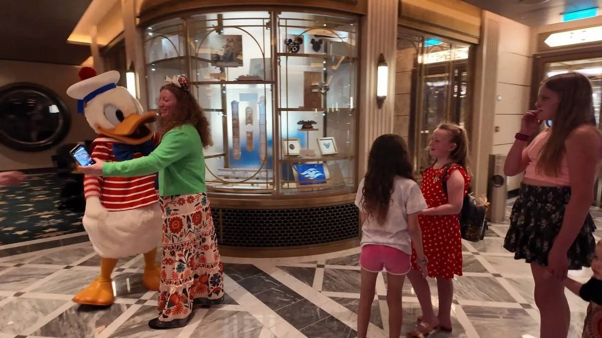 A person wearing a colorful outfit happily hugs a Donald Duck character in a lively indoor setting, while three children watch nearby with smiles. The area features elegant marble flooring and a display case in the background.