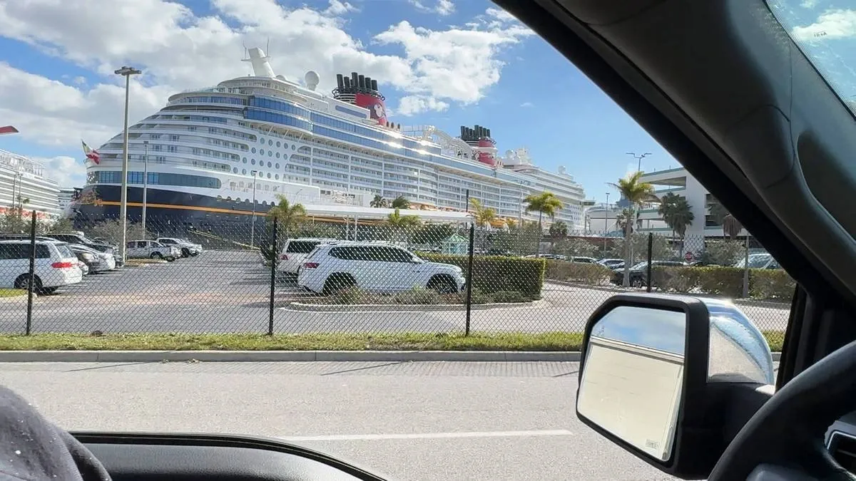 A large cruise ship with distinctive red funnels is docked near a parking lot filled with white cars, viewed from inside a vehicle with part of the side mirror visible. The scene is set under a bright blue sky with scattered clouds.