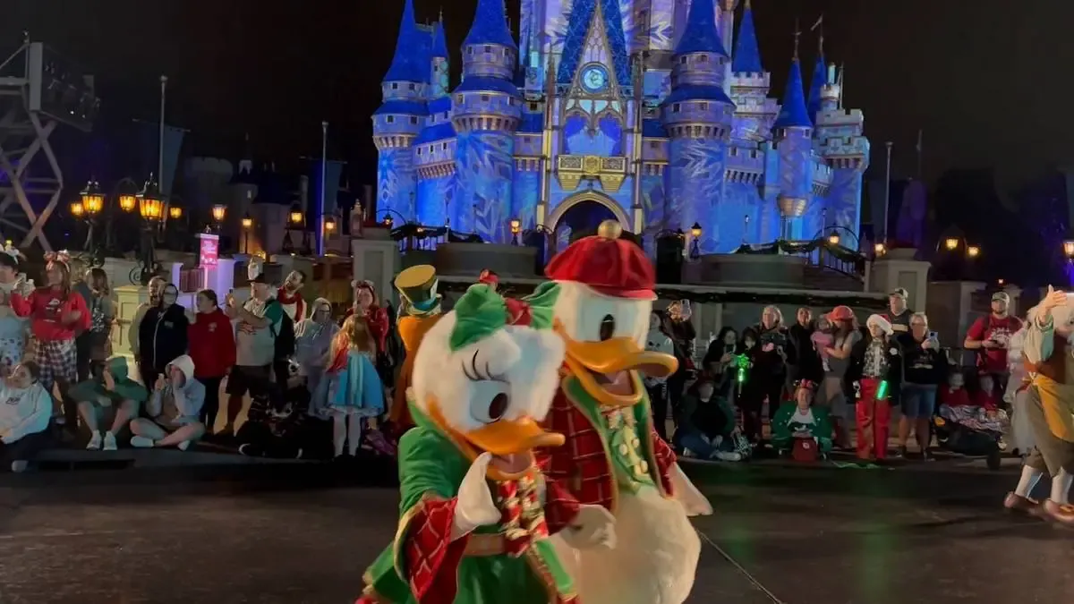 Donald and Daisy Duck, dressed in festive holiday outfits, walk in front of Magic Kingdom castle at night, surrounded by a crowd of onlookers enjoying the scene.