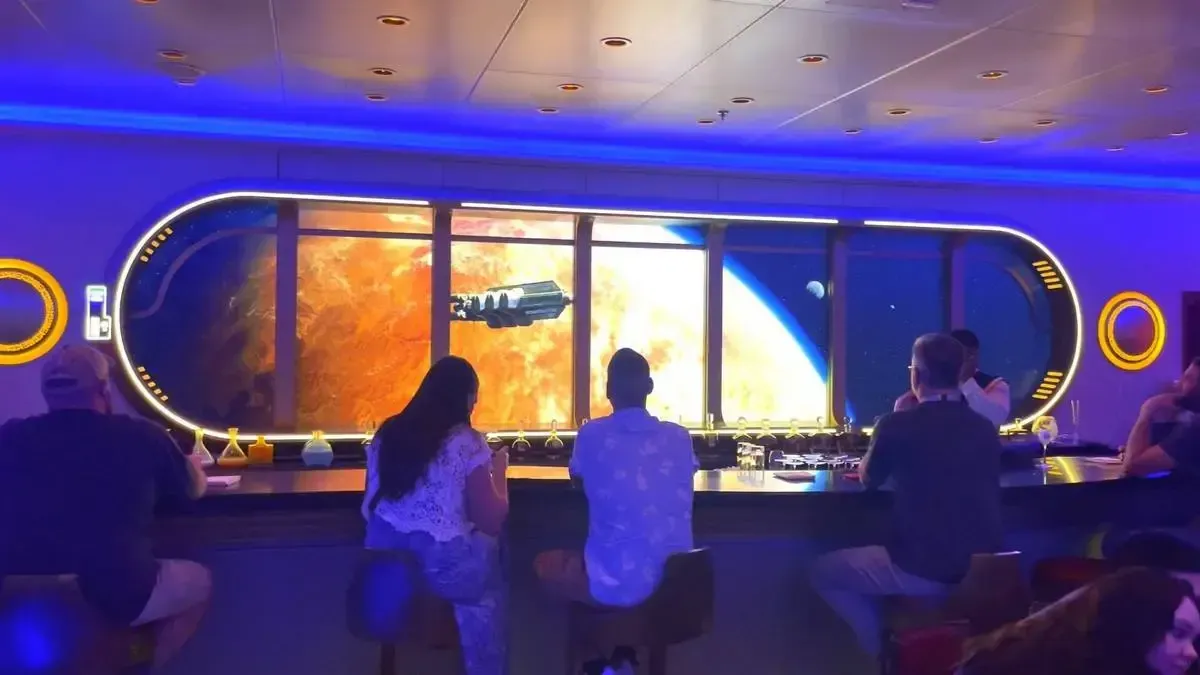 People sit at a futuristic bar with a large screen displaying a space scene, including a planet and a spaceship. The ambient blue lighting enhances the sci-fi atmosphere.
