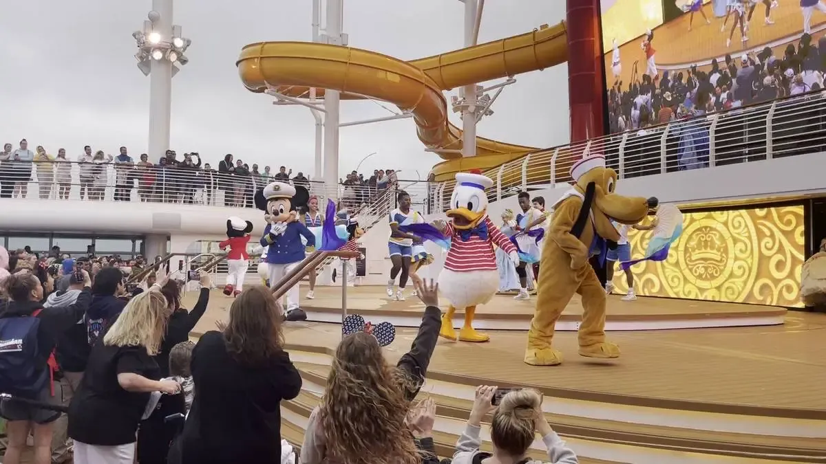 Characters in colorful costumes, including a sailor-themed Mickey Mouse and Donald Duck, entertain a crowd on a ship's deck with a water slide in the background, while excited onlookers cheer and take photos.